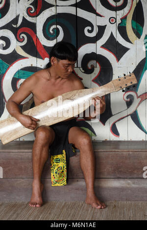 Native Mann spielt traditionelle Musik auf einem elektrischen Sape in der Orang East Langhaus, vor dem Baum des Lebens Wandbild im Sarawak Cultural Village Stockfoto