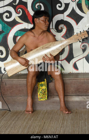 Native Mann spielt traditionelle Musik auf einem elektrischen Sape in der Orang East Langhaus, vor dem Baum des Lebens Wandbild im Sarawak Cultural Village Stockfoto