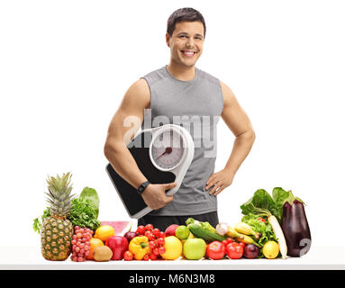 Junge Mann in Sportkleidung Holding eine Waage hinter einem Tisch mit Obst und Gemüse auf weißem Hintergrund gekleidet Stockfoto