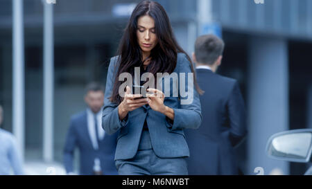 Business Woman im Maßanzug zu Fuß auf der geschäftigen Großstadt Straße im Geschäftsviertel, überprüft Ihr Smartphone. Selbstbewusste Frau auf der Straße. Stockfoto