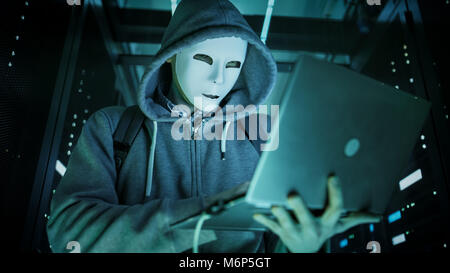 Nahaufnahme einer maskierten Hacker in einem Hoodie steht in der Mitte des Rechenzentrums voll von Rack Servern und Hacking es mit seinem Laptop. Stockfoto