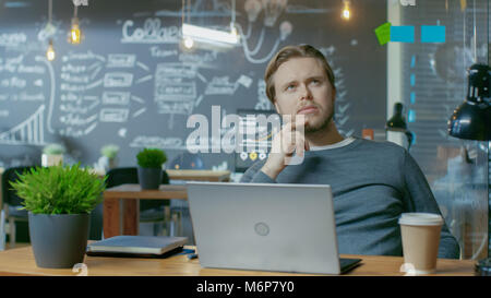 Hübscher junger Mitarbeiter denkt über ein Problem Lösung während der Arbeit auf einem Laptop. Er arbeitet in der Kreativen stilvolle Büro. Stockfoto