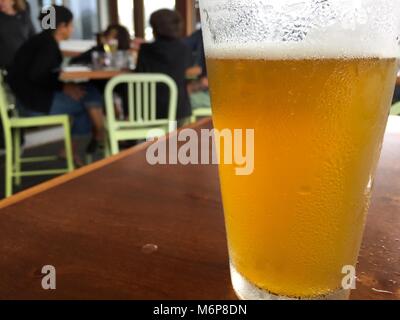 Nahaufnahme Foto von Tall Pint Bier Glas Milchglas mit leichten Lager gegossen sitzen auf Holztisch auf zwangloses Restaurant. Familie genießt Mahlzeit zusammen in Bl Stockfoto