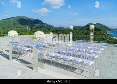 Der Hochzeit, im Freien mit Meerblick und Blick auf die Berge im Hintergrund. Die transparente Stühle für den Gast, den Glaskasten von Blumen, Blumen Stockfoto