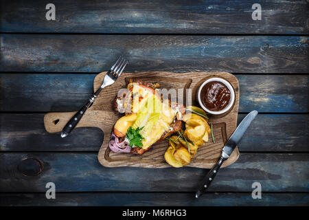 Mit Tomaten, Pilzen und Käse überbacken. Mit Kartoffelecken und BBQ-Soße, mit Besteck und Holz- Hintergrund, Ansicht von oben serviert. Stockfoto