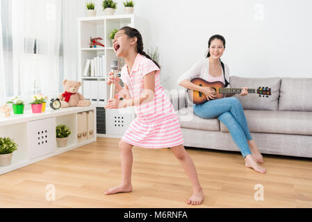 Kleine Schwester hält Ihre persönlichen Konzert im Wohnzimmer. Ihre talentierte Mama hilft ihr die Hintergrundmusik mit der Gitarre auf dem Sofa bei t spielen Stockfoto