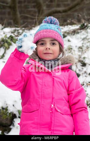 Hübsches kleines Mädchen spielen im Schnee im März winter Holding einen Schneeball trägt einen rosa Fell und blaue Woolly pompom hat. Schnee in Cornwall. Stockfoto