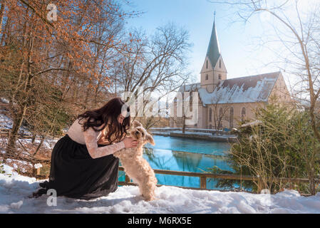 Schöne brünette Frau, elegant gekleidet, ihre havaneser Bichon Hund umfasst, im Schnee am Ufer des Blautopf Frühling, in Blaubeuren, Deutschland. Stockfoto