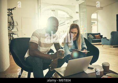 Zwei junge Designer lächelnd und online gemeinsam mit einem Notebook während in einem stilvollen, modernen Büro zu sitzen Stockfoto