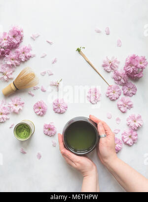 Ansicht von oben von zwei weiblichen Händen halten einer Tasse grünem Matcha Tee, mit Cherry Blumen, Schneebesen und Messlöffel. Stockfoto