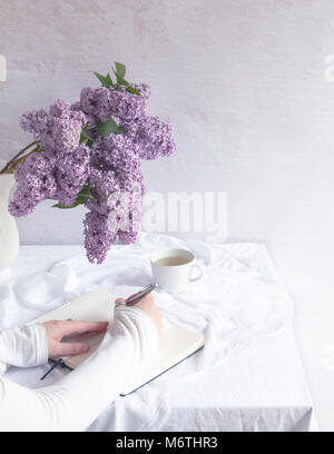 Innen- Szene mit Tisch mit weißer Bettwäsche, Bündel von frischem lila Blumen, Teetasse, Hände auf dem Notebook, eine Hand schreiben Stockfoto