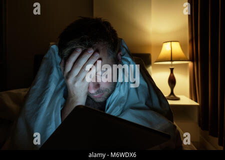 Hübscher junger Mann zu Hause lesen mit Tablet PC liegend auf Bett in der Nacht. Er ist müde und will schlafen Stockfoto