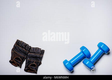Zwei blaue Hanteln liegen auf einem weißen Hintergrund, zusammen mit Sport Handschuhe mit einem Baum Textur. Stockfoto