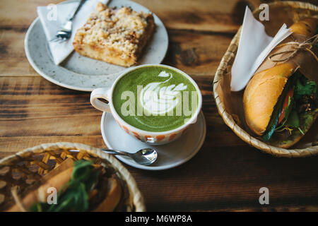 Gesundes Frühstück im Restaurant: Tasse grünen Tee mit Milch, Dessert und Sandwiches mit Gemüse und Kräutern auf Holztisch. Stockfoto
