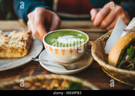 Close-up des Menschen Hand, die eine Tasse grünen Tee mit schönen Muster in Form von weißen Schaum neben Dessert und Sandwich Stockfoto