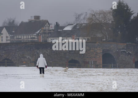 Eine Frau in warme Kleidung geht ihr West Highland Terrier entlang einer öffentlichen Fußweg in einem schneebedeckten Feld in Richtung der Steinstruktur Brücke von Henley Stockfoto