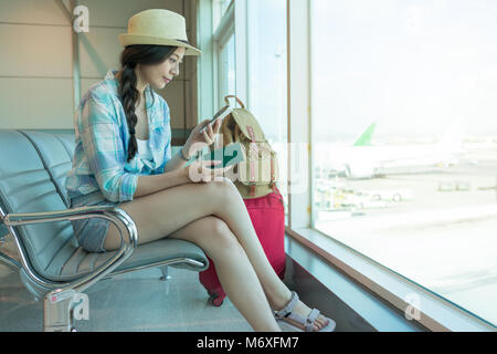 Passagier reisende Frau in Flughafen mit Tablet-Smartphone und halten Ihren Pass. Junge Frau lächelnd Sitzen mit Koffer Trolley im Warten hal Stockfoto