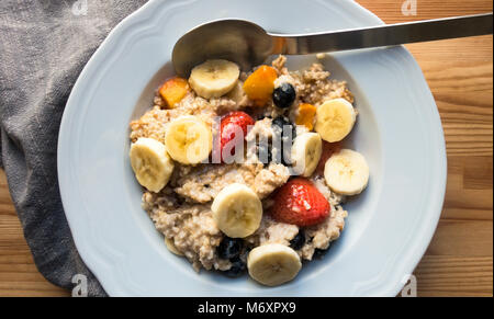 Haferflocken und Obst in einer Schüssel auf einem Holztisch mit einem Löffel und grau Serviette Stockfoto