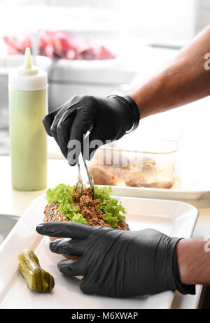 Der Küchenchef bereitet ein gezogener pork Sandwich auf ein gesundes Vollkorn roll Löffelt die Ausschreibung zerfetztes Fleisch auf frischem Salat mit Zange aus Metall Stockfoto