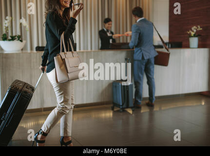 Frau, die Koffer und Gespräch am Handy in der Hotellobby. Reisende Frauen gehen mit ihrem Gepäck im Hotel Flur. Stockfoto