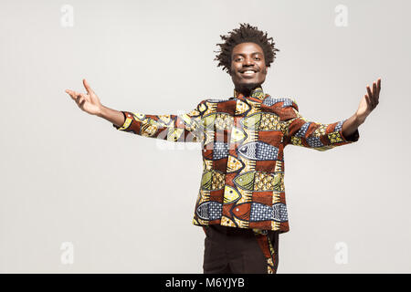 Freundlich afro Mann in traditioneller Kleidung toothy Lächeln, sagen herzlich willkommen. Innen, auf grauem Hintergrund Stockfoto