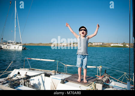 Niedlichen kleinen Jungen in Captain Hut und Sonnenbrille auf Yachtcharter schreit freudig die Hände winken Stockfoto