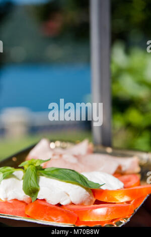 Köstliche italienische Antipasti Teller mit Tomaten, Mozzarella, Basilikum und Prosciutto (Schinken) an einem See für ein romantisches Picknick Stockfoto