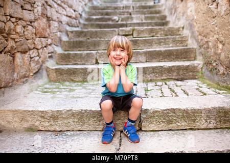 Süß lächelnde Blonde kleine Junge sitzt auf dem alten Kopfsteinpflaster Treppen mit den Händen auf Wangen, im historischen Zentrum von Rovinj