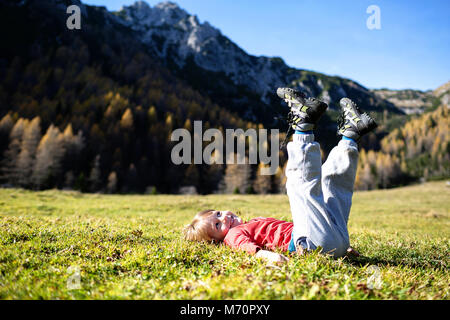 Junge verspielte Blondine western Junge spielt auf dem Gras in der alpinen Landschaft in herbstlichen Farben. Festlegung auf das Gras mit angehobenen Beinen und lächelnd. Stockfoto