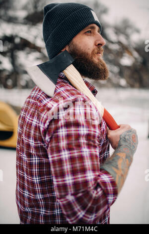 Junge Kerl in Hut und kariertem Hemd mit Tattoo auf der Hand hält Axt in seine Hand im Freien. Stockfoto