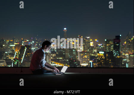 Asiatische Geschäftsmann sitzen und mit dem Laptop über das Stadtbild Hintergrund in der Nacht Zeit, geschäftlichen Erfolg und Technologiekonzept Stockfoto