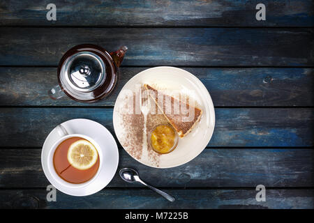 Stück Käsekuchen mit Marmelade und Tee Wasserkocher mit Zitrone auf einem farbigen Hintergrund aus Holz. Ansicht von oben Stockfoto