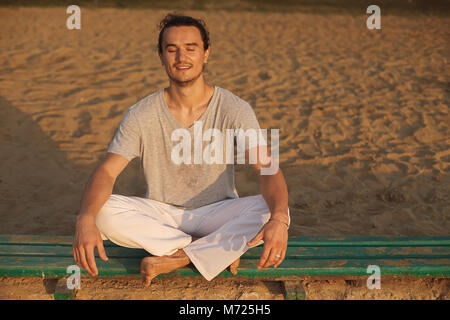 Stattlicher Mann mit geschlossenen Augen lächeln am Strand bei Sonnenaufgang. Portrait von athletischen positive capoeira Mann mit grauen T-Shirt, Ausruhen nach Training". Stockfoto