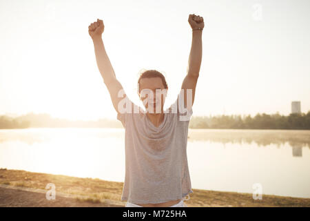 Junge casual sportlich begeistert lächelnde Mann, gekleidet in Grau verschwitzten T-Shirt, mit erhobenen Händen glücklich Training zu beenden. Porträt der Mann in der Nähe der See in Stockfoto