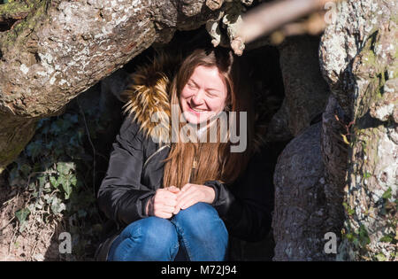 Hübsche junge Frau sitzt draußen unter einem Baum trägt einen Mantel, an einem kalten Tag im Winter mit Sonne im Gesicht. Stockfoto