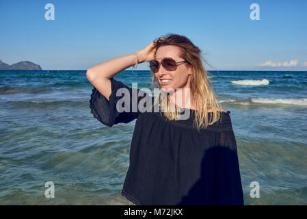 Gerne attraktive blonde Frau eine Sonnenbrille trug am Meer an einem sonnigen Tag vor dem Meer mit ihrem Haar weht im Sea Breeze gri Posing Stockfoto