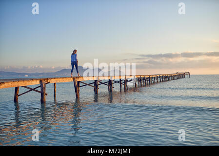 Frau zu Fuß von der Kamera weg entlang einer hölzernen Pier oder Boardwalk bei Sonnenuntergang über einer ruhigen Ozean mit bunten orange Glühen am Himmel Stockfoto