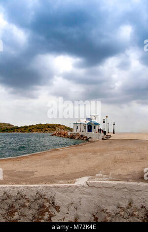 Kleine Kapelle mit weißen Dächern auf Klippe über Meer und Kleine Bucht unter einem dramatischen Himmel auf einer griechischen Insel Stockfoto