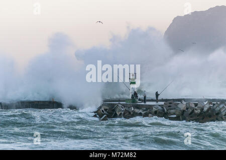 Fischer braving enorme Wellen, die hier zu sehen sind krachend gegen die Wellenbrecher von Kalk Bay Harbor in Kapstadt, Südafrika. Stockfoto