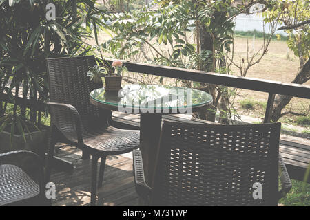 Runde Holz Tisch auf der Terrasse des Hauses. wicker rattan Stuhl auf der Terrasse in der Nähe von Garten zu Hause. entspannten lifestyle Konzept. Stockfoto