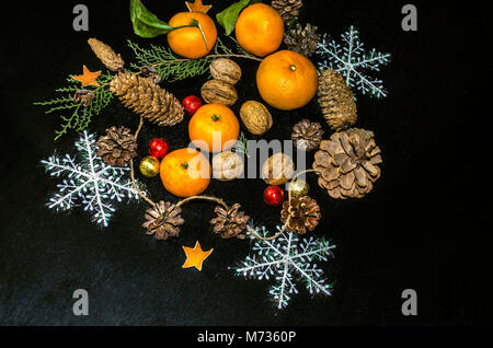 Mandarinen mit Blättern, Zweig der Zypresse, der verschiedenen Nadelbaum Kegel, Nüsse, Schokolade Kugeln mit Schneeflocken auf einem schwarzen Sperrholz Stockfoto
