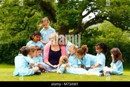 Grundschulkinder, um ihren Lehrer außerhalb unter einer Eiche lesen eine Geschichte gesammelt Stockfoto