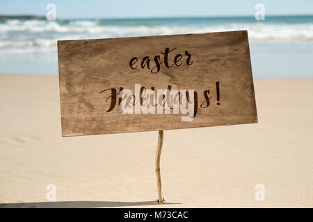 Nahaufnahme einer rustikalen hölzernen Schild mit dem Text Osterferien in Es geschrieben, auf dem Sand von einem ruhigen Strand und das Meer im Hintergrund Stockfoto