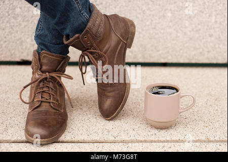 Einen Becher Kaffee auf dem Boden, in der Nähe der Beine von einem Mädchen in Jeans und braune Stiefel Stockfoto