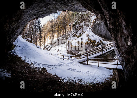 Blick aus dunklen Höhle zu verschneiten Schlucht Baerenschuetzklamm mit Wald und Holz- Brücke im Winter Stockfoto