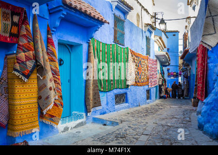 Eine bunte Gasse Szene in die blaue Stadt Chefchaouen, Marokko. Teppiche, Matten, Teppiche auf Anzeige in der gepflasterten Straße mit Menschen in Sicht Stockfoto