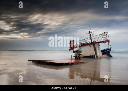 Die gebrochene Schiff zusammen mit dem Meer auf einem sandigen Strand und Sonnenuntergang Dämmerung Himmel. Migrant Boot Stockfoto