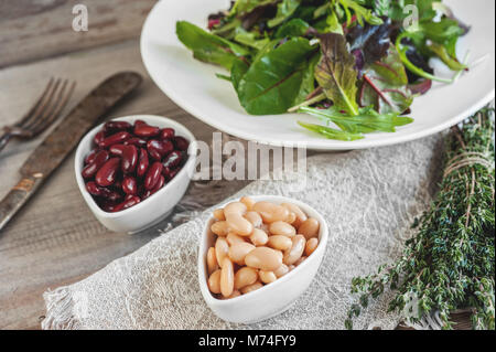 Bohnen sind Weiß und Rot mit großen schwarzen Oliven in Weiß kleine Tassen mit grünen Blättern von Rucola und Lollo Ross für Salat mit Thymian vorbereitet. Verhindern Stockfoto