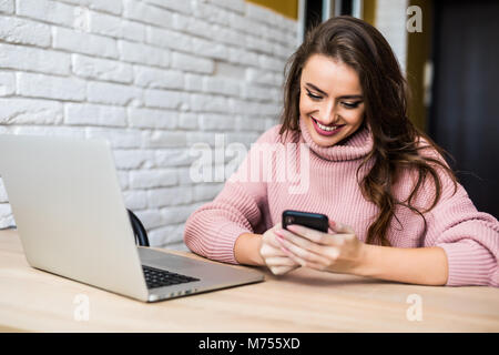 Schöne lächelnde Frau mit ihrem Handy am Tisch mit Laptop im Home Stockfoto