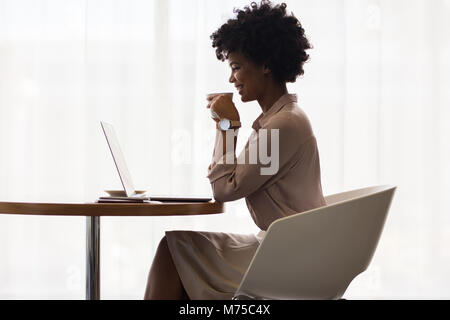 Afrikanische Geschäftsfrau, Lächeln, während mit Laptop und trinken Kaffee im Büro. Seitenansicht der Arbeitnehmer/in während der Arbeit am Laptop. Stockfoto